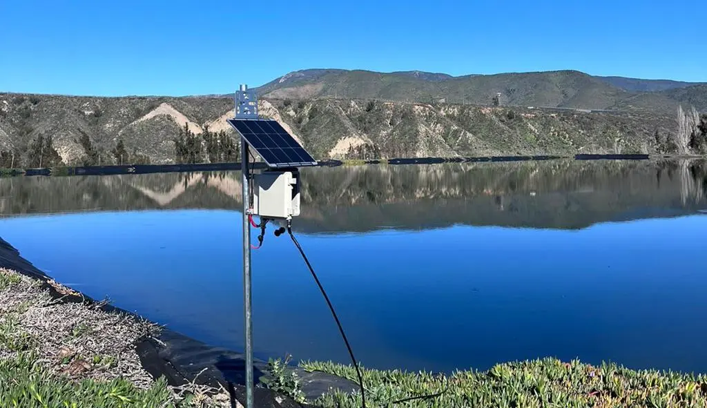 Sensor de nivel con panel solar instalado en la orilla de un tranque, con cerros de fondo